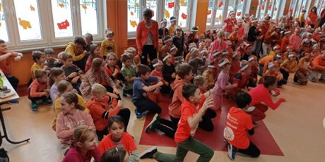 Powiększ grafikę: Duża grupa dzieci ubrane na pomarańczowo, tańczą na korytarzu.