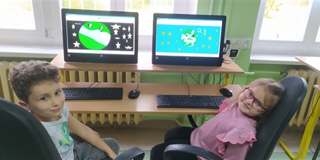 Powiększ grafikę: dwoje dzieci z 1b siedzi przy komputerach i pokazują planetę Ziemię wykonaną w programie Paint