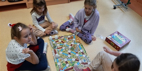 Powiększ grafikę: Dziewczynki siedzą w sali na podłodze, grają w grę planszową.