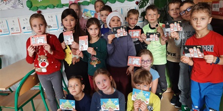 Powiększ grafikę: Dziewczynki I chłopcy stoją,  w sali uśmiechają się. Trzymają w rękach pocztówkę  na pocztówce są Świąteczne życzenia.