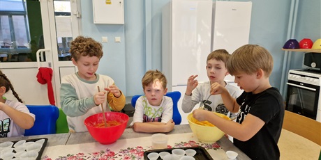 Powiększ grafikę: dzieci siedzą przy stole trzymają miski i wyrabiają w nich ciasto na babeczki i wkładają do foremek