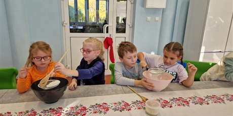 Powiększ grafikę: dzieci siedzą przy stole trzymają miski i wyrabiają w nich ciasto na babeczki