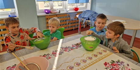 Powiększ grafikę: dzieci siedzą przy stole trzymają miski i wyrabiają w nich ciasto na babeczki