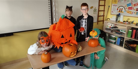 Powiększ grafikę: Dzieci w strojach przebrane za dynie stoją przy tablicy, przed nimi leżą na ławce okazy dyni. 