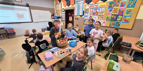Powiększ grafikę: uczniowie siedzą w ławkach, a troje uczniów częstuje dzieci jabłkami z koszyka
