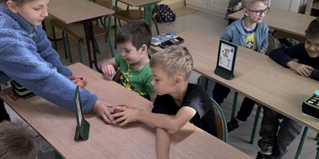 Powiększ grafikę: Dziewczynka w niebieskiej bluzie rozdaje jabłka chłopcom siedzącym w szkolnych ławkach.