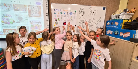Powiększ grafikę: Dziewczynki stoją pod tablicą I wskazują swoją pracę plastyczną
