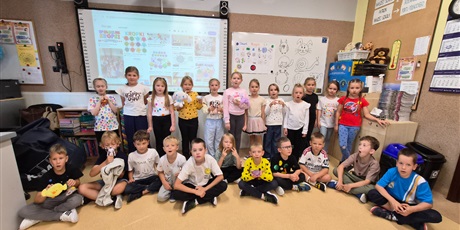 Powiększ grafikę: Dzieci z klasy 1b ubrani w bluzki w kropki stoją pod tablicą ustawione w dwóch rzędach na tle tablicy z obrazkami