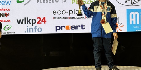 Powiększ grafikę: Dziewczynka stoi na najwyższym stopniu podium z medalem na szyi, z pucharem i dyplomem w rękach.