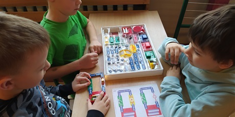 Powiększ grafikę: Troje chłopców siedzi w ławce, przed nimi leżą rysunki obwodów elektrycznych, pudełko z elementami do złożenia, zbudowany układ.