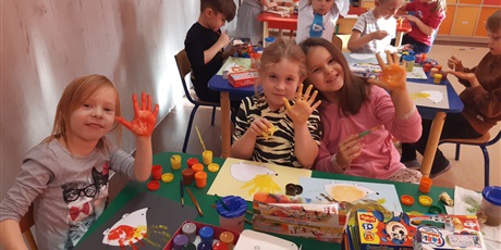 Powiększ grafikę: Dzieci malują swoje dłonie farbami, aby później odbić je na jeżu. Dziewczynki uśmiechają się. 
