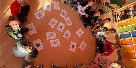 Powiększ grafikę: Dzieci siedzą na podłodze, trzymają się za ręce. W środku kółka znajdują się kolorowanki jakie wykonały.