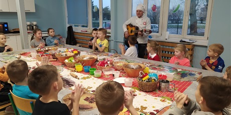 Powiększ grafikę: Dzieci siedzą wokół wielkiego stołu, za nimi stoi pan w czapce kucharza i gra na gitarze.