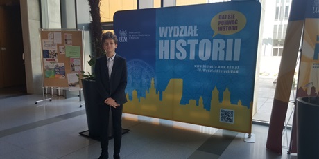 Powiększ grafikę: uczeń stoi przed banerem wydziału historii CH