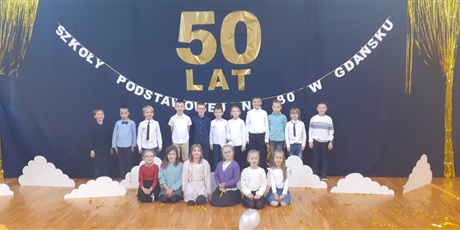 Powiększ grafikę: Uczniowie klasy 1c prezentują się na sali gimnastycznej, w tle dekoracja z napisem 50 lat SP80.