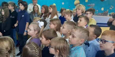 Powiększ grafikę: Dzieci stoją na holu i przygotowują się do odśpiewania pieśni szkoły pt. "Osiemdziesiątka" wraz z całą społecznością szkolną.