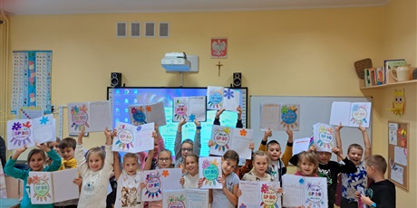Powiększ grafikę: Dzieci stoją przed tablicą, w rękach trzymają zeszyty z kolorowanką.