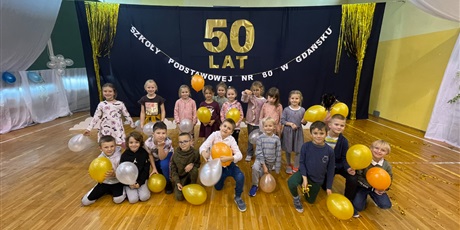 Powiększ grafikę: W pierwszym rzędzie dzieci siedzą lub klęczą, tylny rząd dzieci stoją. W rekach trzymają balony, za nimi napis: 50 lat Szkoły Podstawowej nr 80 w Gdańsku.
