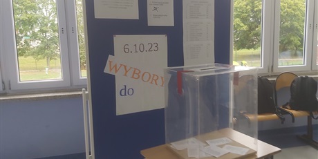 Powiększ grafikę: Przezroczysta urna wyborcza ustawiona na stoliku na tle niebieskiej tablicy z instrukcjami i listami kandydatów do wyborów Samorządu Uczniowskiego. 
