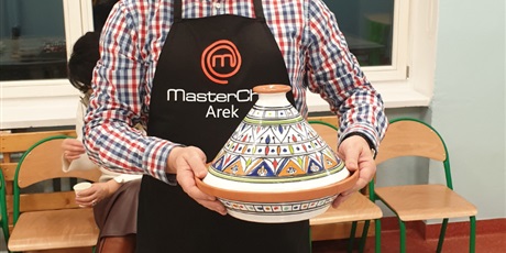 Powiększ grafikę: Po środku mężczyzna w stroju kucharza MasterChef, trzyma w rękach naczynie do gotowania potraw
