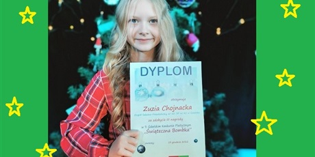 Powiększ grafikę: Dziewczynka trzyma w ręku dyplom za zdobycie III miejsca w Gdańskim Konkursie Plastycznym "Świąteczna Bombka". W tle choinka ozdobiona bombkami.