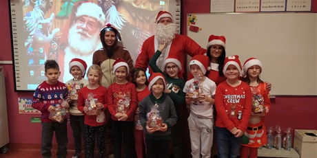 Powiększ grafikę: Grupa dzieci stoi trzymając w rękach paczki z prezentami, za nimi stoi Mikołaj, pani Mikołajowa i renifer.