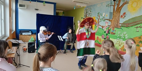 Powiększ grafikę: Pięcioro dzieci stoi tyłem, patrzą na grające osoby na skrzypcach i akordeonie oraz tańczącą postać w wielkim kapeluszu i w trójkolorowym  ponczo
