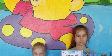 Powiększ grafikę: Dwie dziewczynki stoją na tle ściany z Kubusiem Puchatkiem, w dłoniach trzymają dyplomy i nagrody książkowe.
