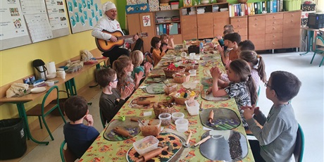 Powiększ grafikę: Wokół stołu siedzą dzieci, słuchają pana ubranego w strój kucharza, grającego na gitarze.