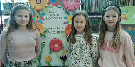 Powiększ grafikę: Trzy dziewczynki stoją, za nimi tablica z kolorowymi kwiatami i napisem: Szkolny konkurs recytatorski "Wiosna daje nadzieje"